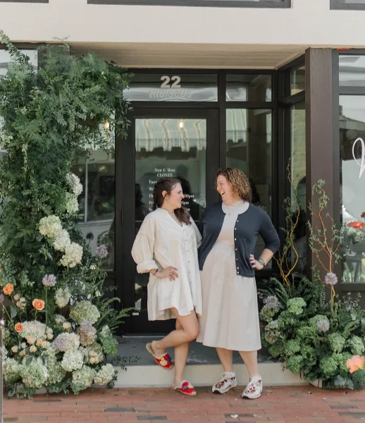 Two women standing in front of the Sweetwood Bakery storefront at 22 Hibiscus St, decorated with floral arrangements.