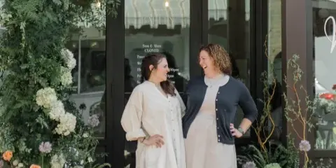 Two women standing in front of the Sweetwood Bakery storefront at 22 Hibiscus St, decorated with floral arrangements.