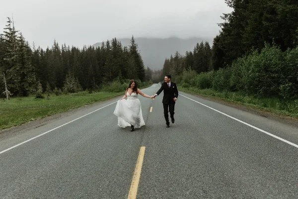 A bride and groom holding hands and walking down a paved road surrounded by evergreen forest and mountains.
