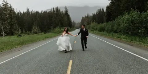 A bride and groom holding hands and walking down a paved road surrounded by evergreen forest and mountains.