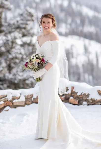 A smiling bride in a white gown and styled updo holding a bouquet in a snowy mountain landscape.