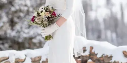 A smiling bride in a white gown and styled updo holding a bouquet in a snowy mountain landscape.