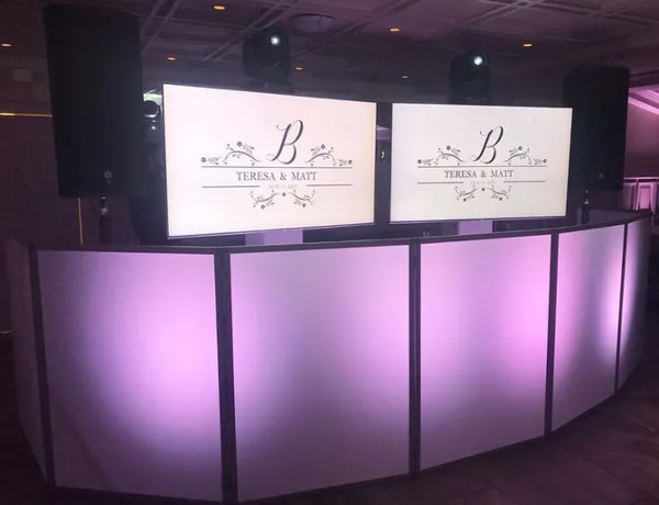 A white DJ booth with purple uplighting and two screens displaying a custom monogram for a wedding couple.