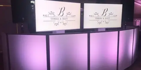 A white DJ booth with purple uplighting and two screens displaying a custom monogram for a wedding couple.
