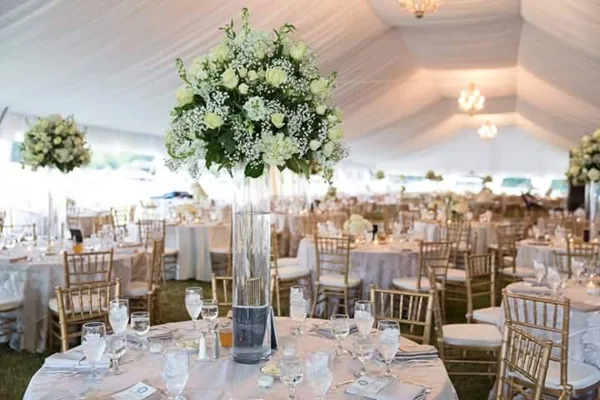 Interior of a white wedding tent with round tables, gold chiavari chairs, white linens, and tall floral centerpieces.