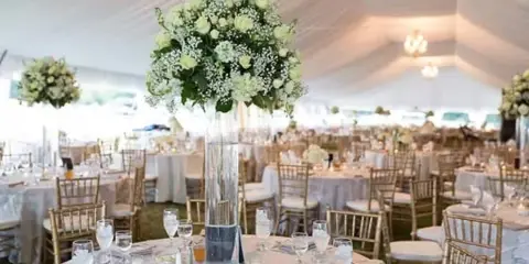 Interior of a white wedding tent with round tables, gold chiavari chairs, white linens, and tall floral centerpieces.
