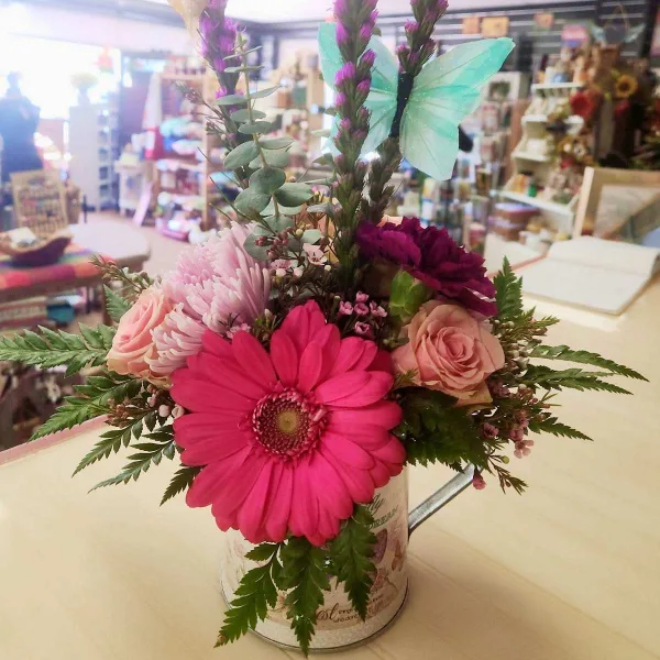 A colorful floral arrangement featuring a large pink gerbera daisy, roses, and a decorative butterfly pick, displayed in a rustic tin container inside a flower shop.