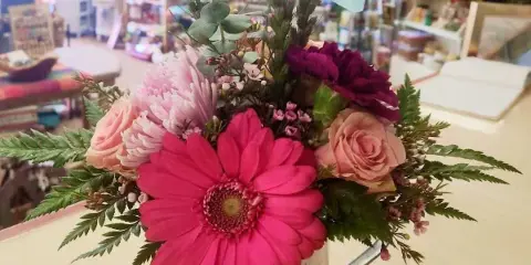 A colorful floral arrangement featuring a large pink gerbera daisy, roses, and a decorative butterfly pick, displayed in a rustic tin container inside a flower shop.