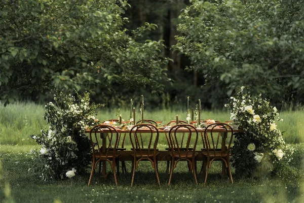 An outdoor dining table set with wooden chairs, candles, and floral arrangements in a grassy field.