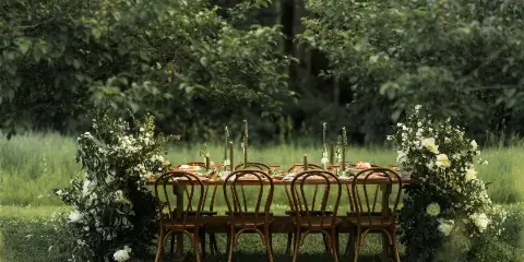 An outdoor dining table set with wooden chairs, candles, and floral arrangements in a grassy field.