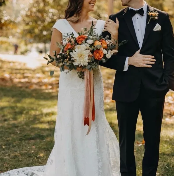 A bride in a white lace gown holding a bouquet of orange and white flowers with a groom in a black tuxedo wearing a matching boutonniere.