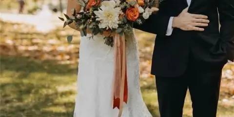 A bride in a white lace gown holding a bouquet of orange and white flowers with a groom in a black tuxedo wearing a matching boutonniere.