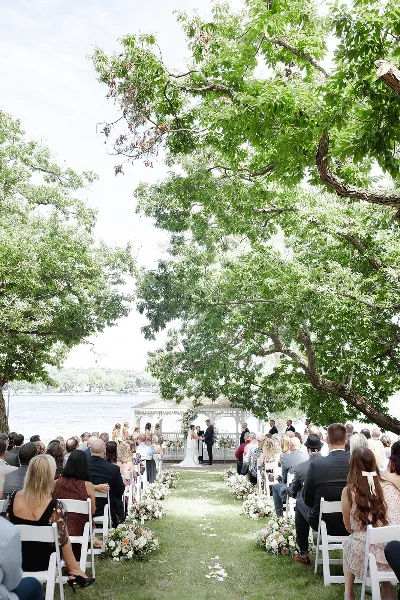A wedding ceremony taking place outdoors under large trees with a groom and bride at the altar and guests seated in white chairs.