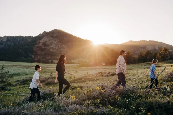 A family of four walking through a grassy field with wildflowers at sunset, with mountains in the background.