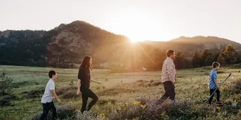 A family of four walking through a grassy field with wildflowers at sunset, with mountains in the background.