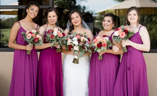 Bride and bridesmaids in purple dresses holding colorful wedding bouquets with roses, dahlias, and eucalyptus.