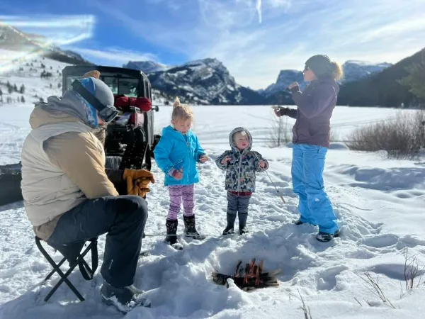 A group of people, including children, enjoying a private outdoor activity with a small fire in a snowy mountain landscape.