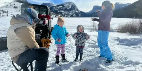 A group of people, including children, enjoying a private outdoor activity with a small fire in a snowy mountain landscape.