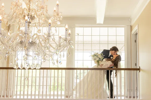 A bride and groom sharing a kiss on an indoor balcony with a large crystal chandelier and white railings.