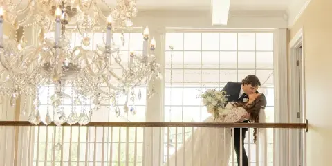 A bride and groom sharing a kiss on an indoor balcony with a large crystal chandelier and white railings.