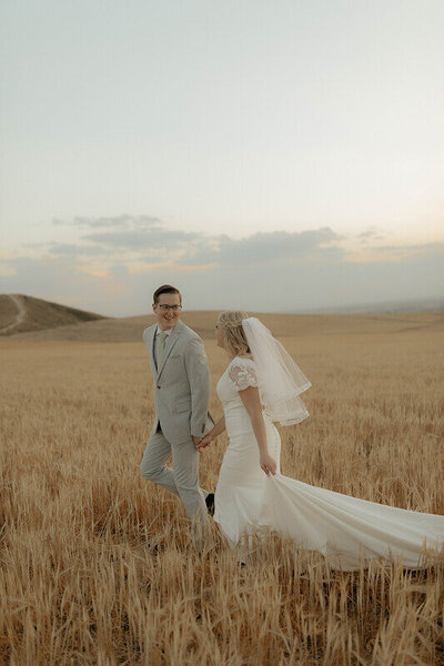 A bride and groom holding hands and walking through a golden wheat field at sunset.