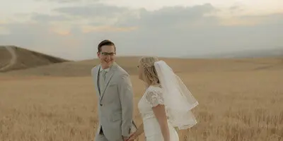 A bride and groom holding hands and walking through a golden wheat field at sunset.