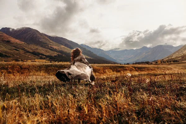 A woman in a flowing dress standing in a grassy field with mountains in the background.