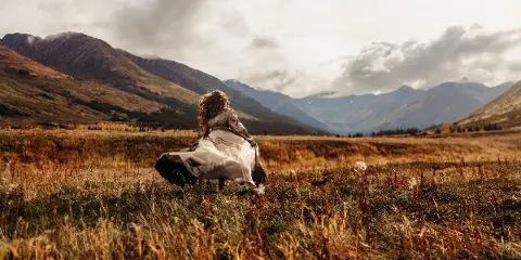 A woman in a flowing dress standing in a grassy field with mountains in the background.