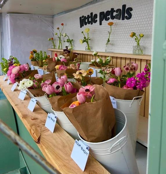 A flower farm stand displaying colorful bouquets in white buckets with the 'little petals' logo on the wall.