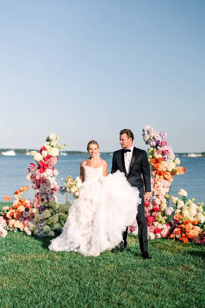 A bride in a voluminous white gown and a groom in a black tuxedo standing on a grassy lawn in front of a vibrant floral arch with a body of water in the background.