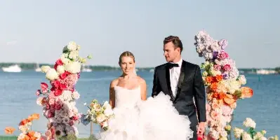 A bride in a voluminous white gown and a groom in a black tuxedo standing on a grassy lawn in front of a vibrant floral arch with a body of water in the background.