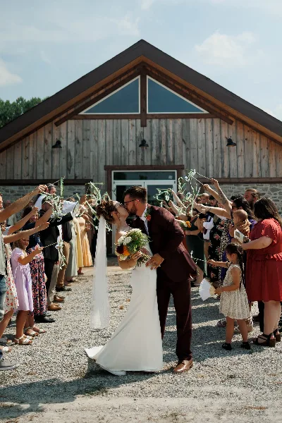 A bride and groom kiss while walking down a gravel aisle lined with guests, with a large wooden barn structure in the background.