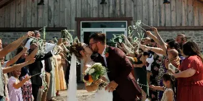 A bride and groom kiss while walking down a gravel aisle lined with guests, with a large wooden barn structure in the background.