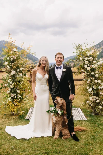 A bride and groom posing with a dog in front of floral wedding arches with a mountain background.