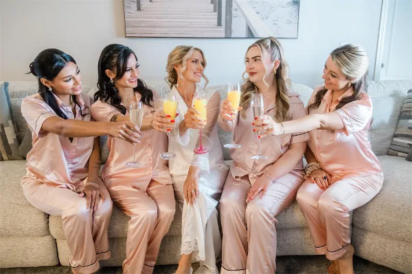 A bride and four bridesmaids in matching pink silk pajamas sitting on a sofa and toasting with champagne flutes.