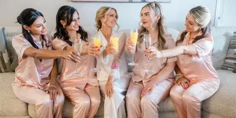 A bride and four bridesmaids in matching pink silk pajamas sitting on a sofa and toasting with champagne flutes.