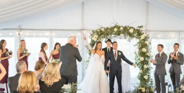 A wedding officiant stands at a floral arch with a bride and groom during a ceremony inside a white tent.