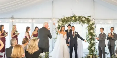 A wedding officiant stands at a floral arch with a bride and groom during a ceremony inside a white tent.