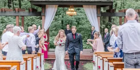 A bride and groom walking down the aisle at an outdoor wedding ceremony under a wooden pergola.