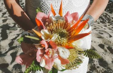 A bride holding a tropical wedding bouquet with pink and orange flowers on a sandy beach.