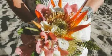 A bride holding a tropical wedding bouquet with pink and orange flowers on a sandy beach.