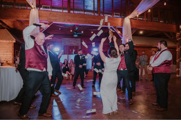 A wedding DJ in a red vest holds a microphone and dances with the bride and groom on a dance floor while guests throw money in the air.