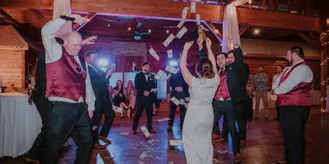A wedding DJ in a red vest holds a microphone and dances with the bride and groom on a dance floor while guests throw money in the air.