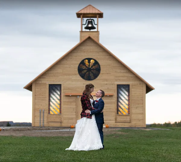A bride and groom standing in front of a wooden chapel with a bell tower and stained glass windows.