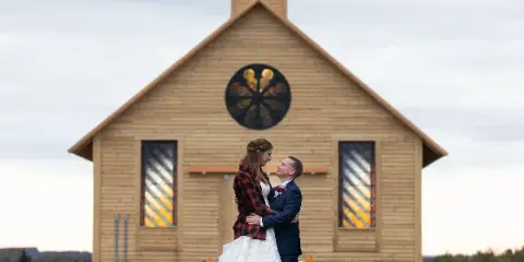 A bride and groom standing in front of a wooden chapel with a bell tower and stained glass windows.