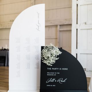 A black and white wedding seating chart and welcome sign with a small floral arrangement displayed in a venue.