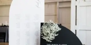 A black and white wedding seating chart and welcome sign with a small floral arrangement displayed in a venue.
