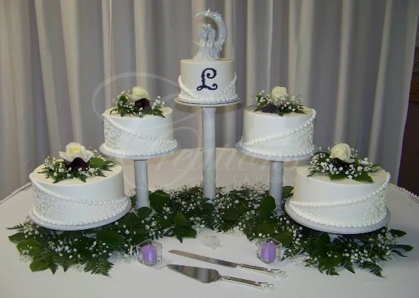 A five-tier white wedding cake decorated with fresh flowers and a bride and groom topper, displayed on a table with greenery.