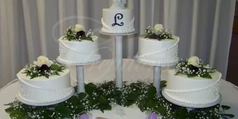 A five-tier white wedding cake decorated with fresh flowers and a bride and groom topper, displayed on a table with greenery.