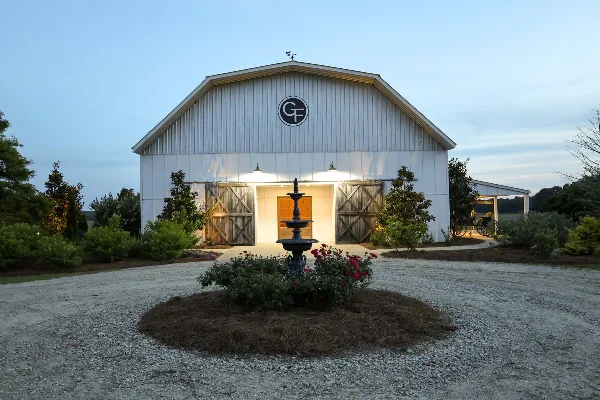 Exterior view of a white barn event venue with a fountain and gravel driveway at dusk.